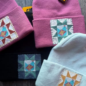 May include: A collection of beanies in various colors, including pink, black, and light blue. Each beanie features a square patch with a bird design in a quilt-like pattern. The beanies have a folded brim.