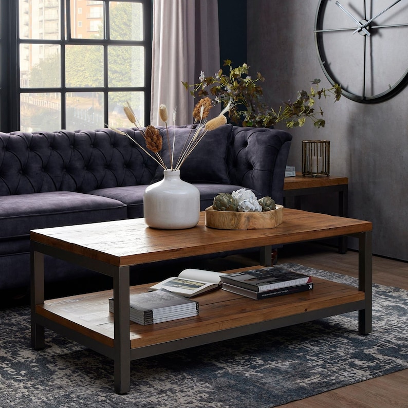 May include: A rustic wooden coffee table with a metal frame and a lower shelf. The table has a distressed wood finish and is decorated with a vase of dried flowers, a bowl of decorative items, and a stack of books.