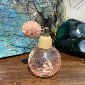 May include: A clear glass atomizer bottle with a pink bulb atomizer and a cream colored top. The bottle has a textured surface and is filled with a clear liquid.
