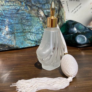 May include: A frosted glass perfume bottle with a gold top and a white tassel. The bottle has a decorative, swirling design.