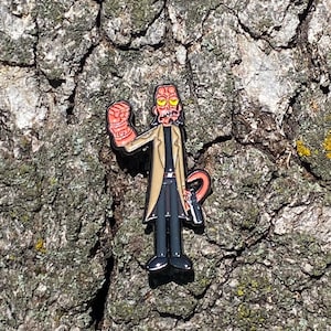 May include: A black and red enamel pin of a cartoon character with a red head, black eyes, and a black beard. The character is wearing a tan trench coat and black pants. The character is holding a gun in their right hand and has a red fist in their left hand.