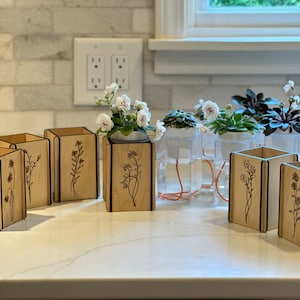 Peut inclure: Un ensemble de six pots de plantes en bois avec des motifs floraux. Les pots sont disposés sur un comptoir blanc. Les pots sont en bois clair et ont une teinte brun foncé. Les motifs floraux sont découpés au laser dans le bois.