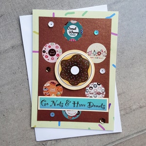 May include: A light green card with a brown background featuring a chocolate donut with sprinkles. The card has a variety of donut-themed stickers with text that reads "Donut Worry", "Treat Yourself", "Sweet Donuts", and "Happy Birthday!" The card also has the text "Go Nuts & Have Donuts" in a blue banner.