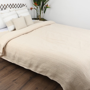 May include: A cream-colored waffle weave throw blanket with fringe on a bed with white pillows. The blanket is made of cotton and is soft and absorbent.