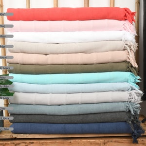 May include: A stack of 12 cotton throw blankets in various colors, including red, pink, white, beige, khaki green, mint, baby blue, light gray, gray, dark gray, navy blue, and black. The blankets are folded and stacked on a wooden shelf.