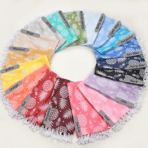 May include: A selection of beach towels in various colours, each featuring a pineapple pattern. The towels are arranged in a circle, with the colours ranging from yellow to blue.
