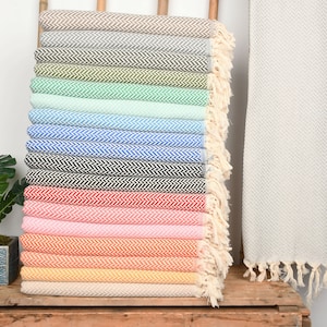 Puede incluir: Una pila de 18 toallas turcas coloridas con un patrón de chevron. Las toallas están dobladas y apiladas en un arcoíris de colores, incluyendo beige, gris, verde, menta, azul claro, azul, negro, rojo, rosa, naranja, melocotón, amarillo y crema. Las toallas tienen flecos en los extremos.