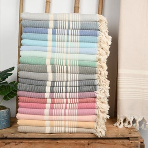 May include: A stack of colorful Turkish towels in various shades, including gray, blue, green, and pink. Each towel features a striped design and fringed edges. A rolled beige towel is on the right.