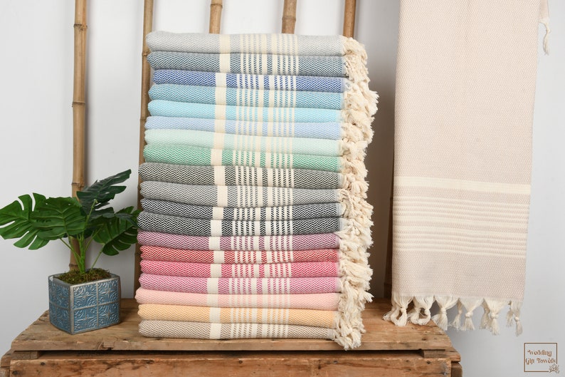 May include: A stack of colorful Turkish towels in various shades, including gray, blue, green, and pink. Each towel features a striped design and fringed edges. A rolled beige towel is on the right.