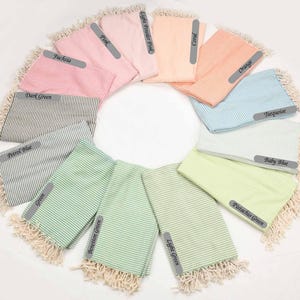 May include: A variety of striped Turkish towels in different colors, including pink, light green, turquoise, and baby blue. The towels are arranged in a circle with the color names written on each towel.
