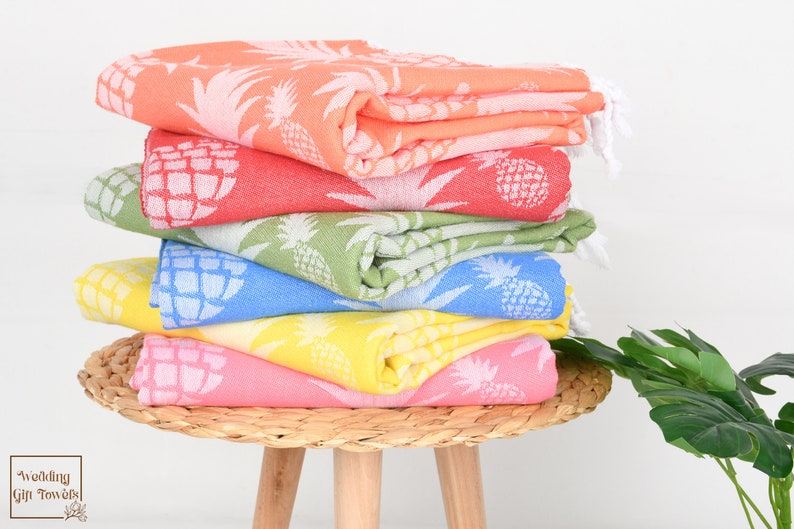 May include: Stack of six colorful Turkish cotton beach towels with pineapple pattern.  Towels are in shades of coral, light green, light blue, yellow, and pink.  Each towel features a white pineapple design.  The towels are neatly folded and stacked on a round woven basket on a small wooden stool. Text on image reads: 'Wedding Gift Towels'.
