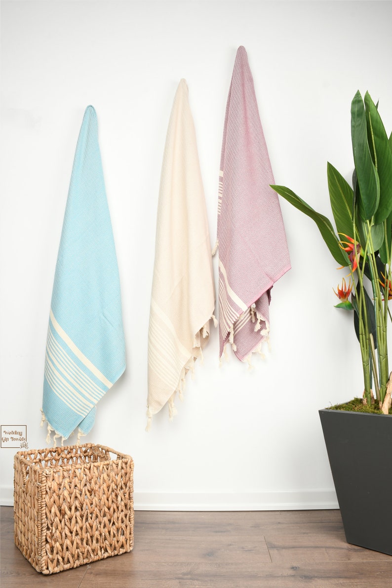May include: Three Turkish towels in light blue, cream, and mauve hang against a white wall. Each towel has white stripes and fringe. A woven basket sits on the floor, and a potted plant is to the right.