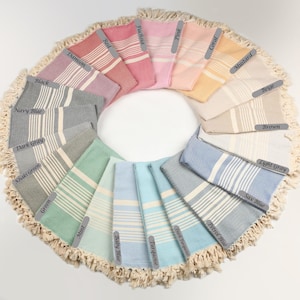 May include: A circular arrangement of Turkish towels in various colors, including navy blue, pink, and turquoise. Each towel features a striped design and is labeled with its color name. The towels are displayed on a white surface, with a fringed border.