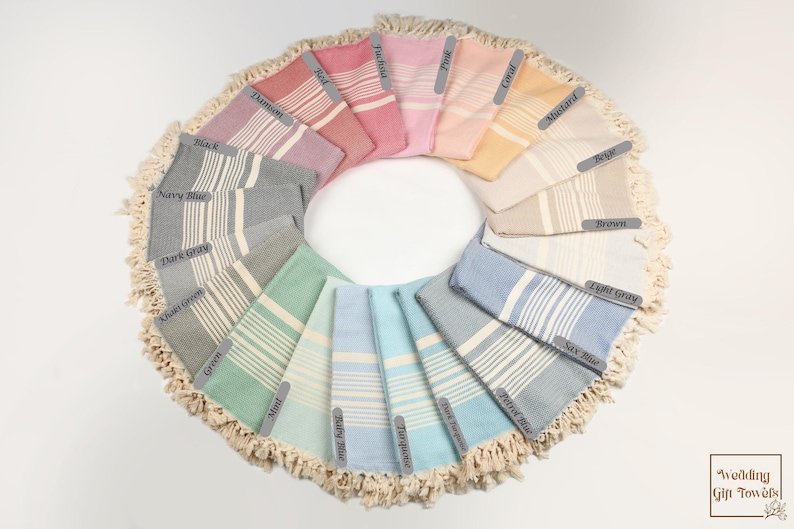 May include: A circular arrangement of Turkish towels in various colors, including navy blue, pink, and turquoise. Each towel features a striped design and is labeled with its color name. The towels are displayed on a white surface, with a fringed border.