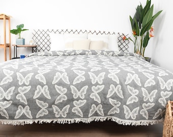 Puede incluir: Una manta gris y blanca con un patrón de mariposas. La manta tiene un borde de flecos y está doblada en una cama con almohadas blancas.