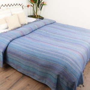May include: A blue and purple striped throw blanket with a fringe border. The blanket is folded on a bed with white pillows.