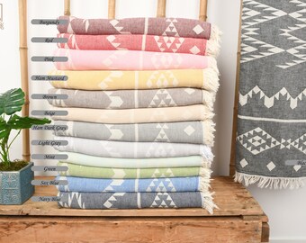 May include: A stack of 12 Turkish towels in various colors, including burgundy, pink, yellow, brown, beige, gray, mint, green, blue, and navy. The towels have a geometric pattern and are fringed on the ends. The towels are displayed on a wooden shelf with a green plant in a pot to the left.