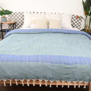 May include: A blue and green patterned throw blanket with white tassels. The blanket is folded on a bed with white pillows.