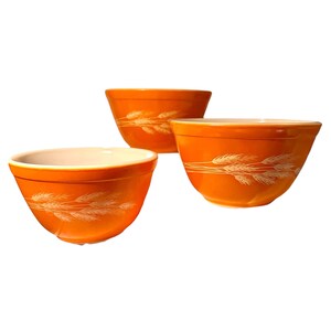 Könnte beinhalten: Drei orangefarbene Keramikschalen mit einem weißen Weizenähren-Design. Die Schalen haben eine glänzende Oberfläche und einen weißen Innenraum. Die Schalen sind unterschiedlich groß und in einer Gruppe angeordnet.