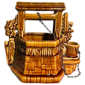 Pode incluir: Um vaso de cerâmica castanha em forma de poço dos desejos com padrão de tijolo, um pequeno balde e uma corrente. A palavra "WISHING" está em relevo na frente. Estética rústica e vintage.