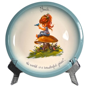 May include: A decorative plate with a light blue rim and a central illustration. The illustration depicts a cartoon girl with red hair sitting on a mushroom, holding a blue bird. The plate has the text "Smile... the world is a wonderful place."