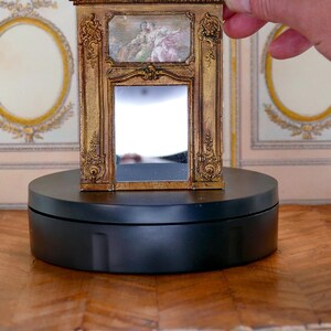 May include: A miniature gold-framed mirror with a decorative design and a small painting inside. The mirror is on a black circular base.