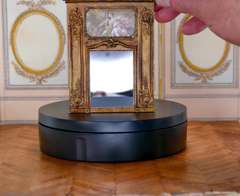 May include: A miniature gold-framed mirror with a decorative design and a small painting inside. The mirror is on a black circular base.