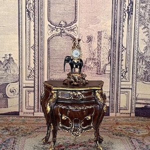 May include: A miniature antique-style table with a dark brown finish and gold accents. A small clock sits atop a black elephant figurine on the table. The background features a vintage-style wall with a door design.