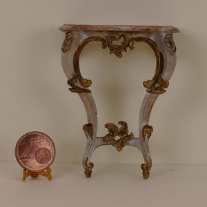 Può includere: Un tavolino da console in miniatura dipinto di bianco e oro con un piano in marmo. Il tavolo presenta volute ornate e un delicato motivo floreale. Il tavolo è circa delle dimensioni di una moneta da due euro.