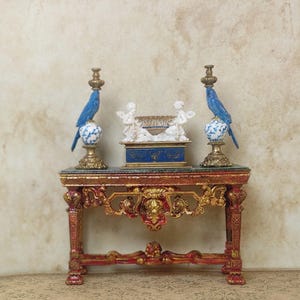 May include: A miniature antique table with ornate red and gold detailing. Two blue bird candle holders flank a blue and white box with cherub figurines. The table has a marble-like top.