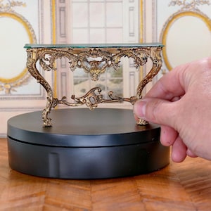 Puede incluir: Una mesa consola en miniatura de color dorado con una tapa de vidrio, que se asienta sobre una base circular negra. La mesa tiene intrincados detalles de volutas y un estilo rococó.