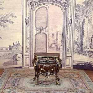 Peut inclure: Une petite table de chevet en bois de style antique avec des accents dorés ornés. La table de chevet a un dessus incurvé et un seul tiroir. Elle repose sur quatre pieds incurvés. Le fond présente un papier peint vintage et un tapis floral.