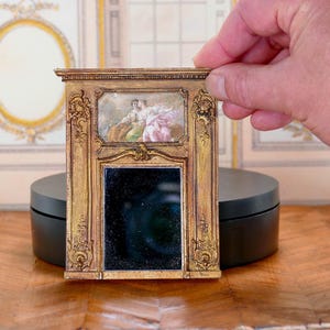 May include: A miniature gold-framed mirror with a black reflective surface. The frame has intricate carvings and a small painting of two figures in the top section.