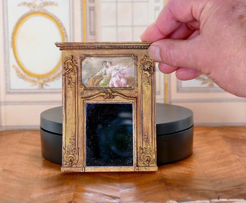 May include: A miniature gold-framed mirror with a black reflective surface. The frame has intricate carvings and a small painting of two figures in the top section.