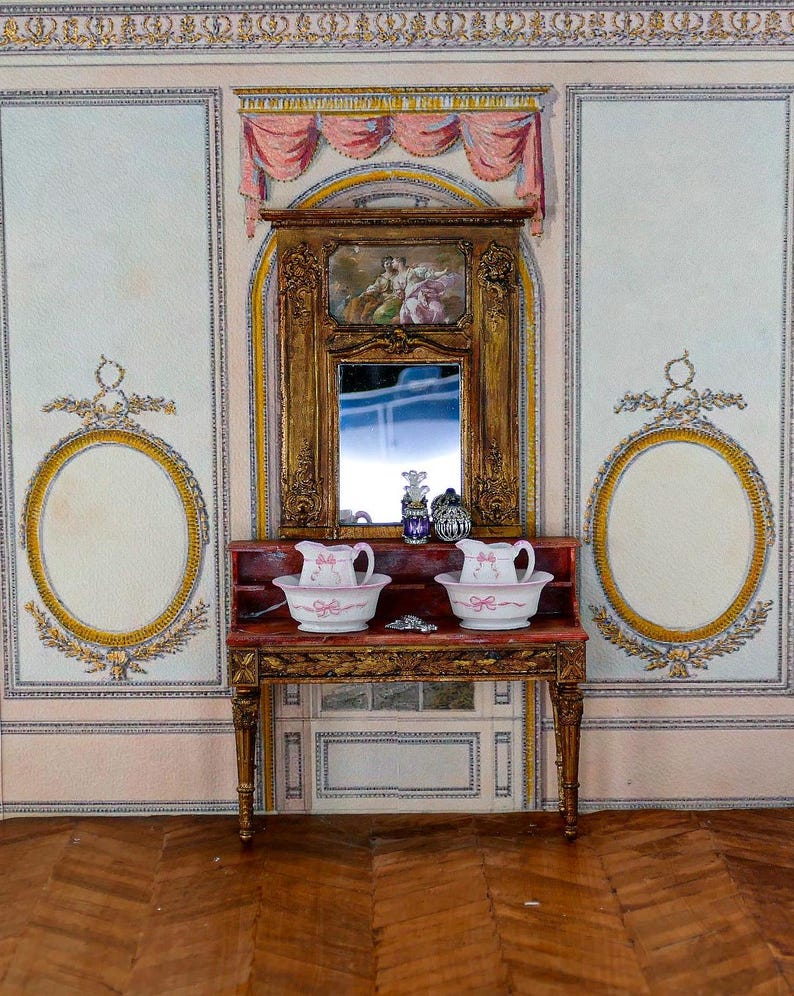 May include: A miniature dollhouse bathroom with a white and gold patterned wallpaper, a wooden vanity with two porcelain pitchers and basins, and a framed mirror with a painted scene above it.