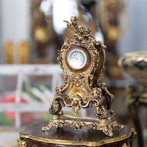 Puede incluir: Un reloj decorativo de color dorado con un diseño ornamentado y una esfera blanca. El reloj está colocado sobre una base de madera.