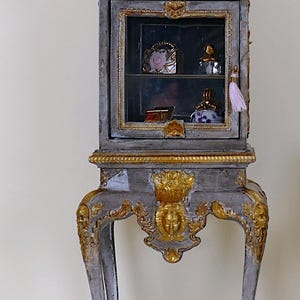 Könnte beinhalten: Miniatur-Schrank in Grau und Gold mit Glastür, Zierdetails und geschwungenen Beinen. Der Schrank ist mit kleinen Dekorationsartikeln gefüllt. Eine 2-Euro-Cent-Münze befindet sich neben dem Schrank.