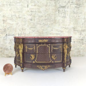 Puede incluir: Una cómoda de madera de estilo antiguo en miniatura con una tapa de mármol rojo, detalles dorados y dos cajones. La cómoda está decorada con tallas intrincadas y tiene un frente curvo.