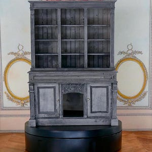 Peut inclure: Un meuble gris en bois avec des étagères vitrées au-dessus d'une partie inférieure avec des portes et une ouverture centrale. Le meuble est posé sur une base noire, avec un fond décoratif.