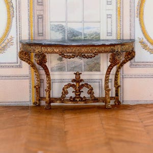 Puede incluir: Una consola de madera dorada con una tapa de mármol negro y tallas ornamentadas. La consola tiene una tapa curva y cuatro patas con detalles intrincados.