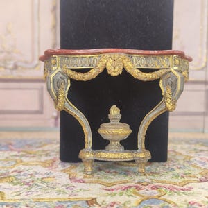 Puede incluir: Una mesa consola miniatura adornada con una tapa de mármol rojo, detalles dorados y tallas intrincadas. La mesa está hecha de madera y tiene un diseño delicado y elegante.