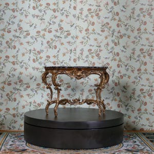 Könnte beinhalten: Ein verzierter Konsolentisch in Gold mit einer schwarzen Marmorplatte steht auf einem schwarzen Sockel. Der Tisch hat ein filigranes, kunstvolles Design mit geschwungenen Details. Der Tisch ist vor einem Hintergrund mit floralem Muster auf der Tapete platziert.