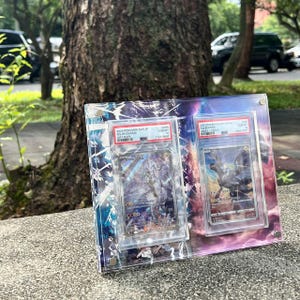 K&ouml;nnte beinhalten: Ein klarer Acryl-Displayrahmen mit zwei bewerteten Pok&eacute;mon-Sammelkarten. Der Rahmen hat einen farbenfrohen, abstrakten Hintergrund mit blauen, lila und wei&szlig;en Akzenten. Die Karten sind in Schutzh&uuml;llen eingeschlossen, mit sichtbarem Text und Bewertungsinformationen.