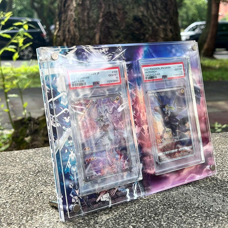 K&ouml;nnte beinhalten: Eine transparente Acryl-Vitrine mit zwei bewerteten Pok&eacute;mon-Sammelkarten. Die Karten sind in Schutzh&uuml;llen eingeschlossen und zeigen farbenfrohe Kunstwerke. Die Vitrine hat einen dekorativen Hintergrund mit einem lila und blauen Design und wird von einem Metallst&auml;nder gehalten.