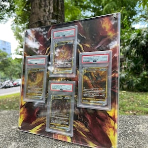 Puede incluir: Una vitrina de acr&iacute;lico transparente con cinco cartas coleccionables de Pok&eacute;mon calificadas. El fondo presenta un dise&ntilde;o abstracto ardiente en naranja y amarillo. Las cartas est&aacute;n en protectores, con informaci&oacute;n de calificaci&oacute;n visible.