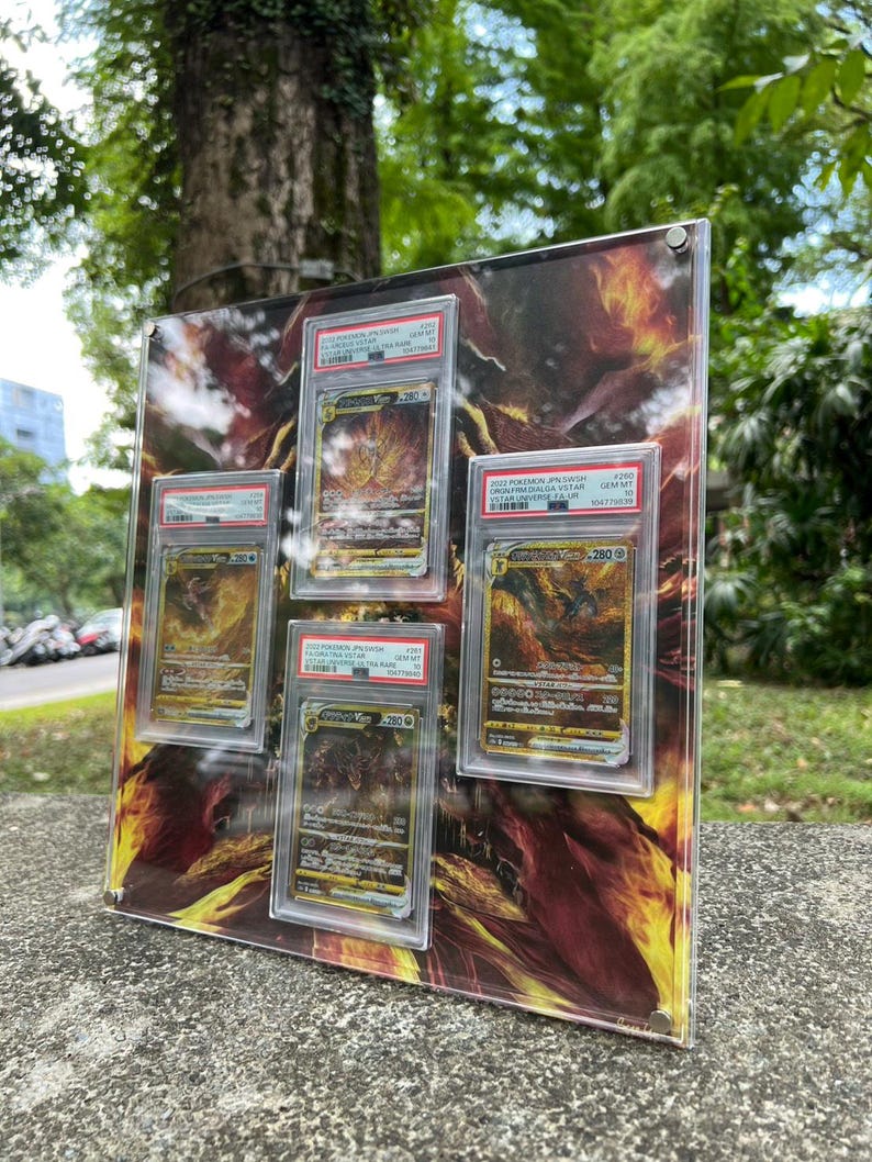 Puede incluir: Una vitrina de acr&iacute;lico transparente con cinco cartas coleccionables de Pok&eacute;mon calificadas. El fondo presenta un dise&ntilde;o abstracto ardiente en naranja y amarillo. Las cartas est&aacute;n en protectores, con informaci&oacute;n de calificaci&oacute;n visible.