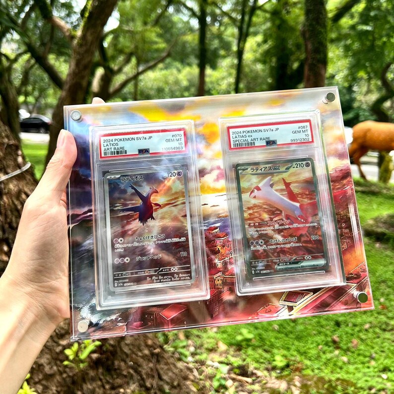 May include: A clear acrylic display case holding two Pok&eacute;mon trading cards. The cards feature images of Latios and Latias, with Japanese text and the text "2024 POKEMON SV7 JP" and "SPECIAL ART RARE". The cards are graded and encased in protective holders.