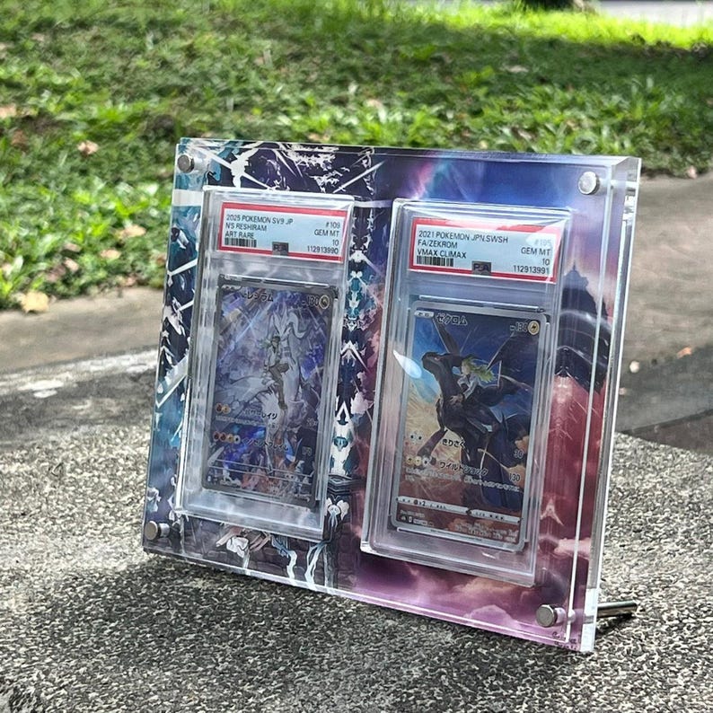 K&ouml;nnte beinhalten: Ein klarer Acryl-Displayrahmen mit zwei Pok&eacute;mon-Sammelkarten. Die Karten befinden sich in Schutzh&uuml;llen, mit japanischem Text. Der Rahmen hat einen farbenfrohen, abstrakten Hintergrund und einen Metallst&auml;nder.
