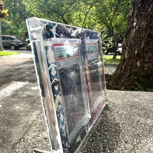 K&ouml;nnte beinhalten: Eine transparente Acryl-Vitrine mit mehreren Sammelkarten. Das Geh&auml;use hat einen dekorativen Rand mit einem blau-wei&szlig;en Muster. Die Karten sind im Inneren des Geh&auml;uses mit Textetiketten sichtbar. Die Anzeige wird auf einer Betonoberfl&auml;che abgest&uuml;tzt.
