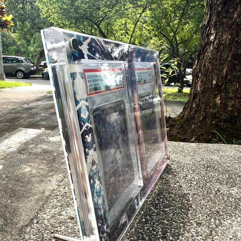 K&ouml;nnte beinhalten: Eine transparente Acryl-Vitrine mit mehreren Sammelkarten. Das Geh&auml;use hat einen dekorativen Rand mit einem blau-wei&szlig;en Muster. Die Karten sind im Inneren des Geh&auml;uses mit Textetiketten sichtbar. Die Anzeige wird auf einer Betonoberfl&auml;che abgest&uuml;tzt.